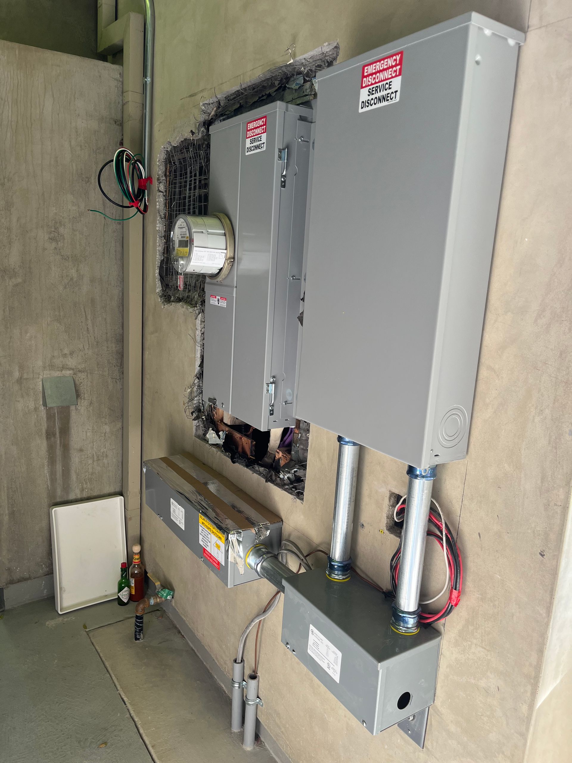 A utility wall featuring electrical meters, circuit breaker boxes, and conduit wiring mounted on a textured gray wall.