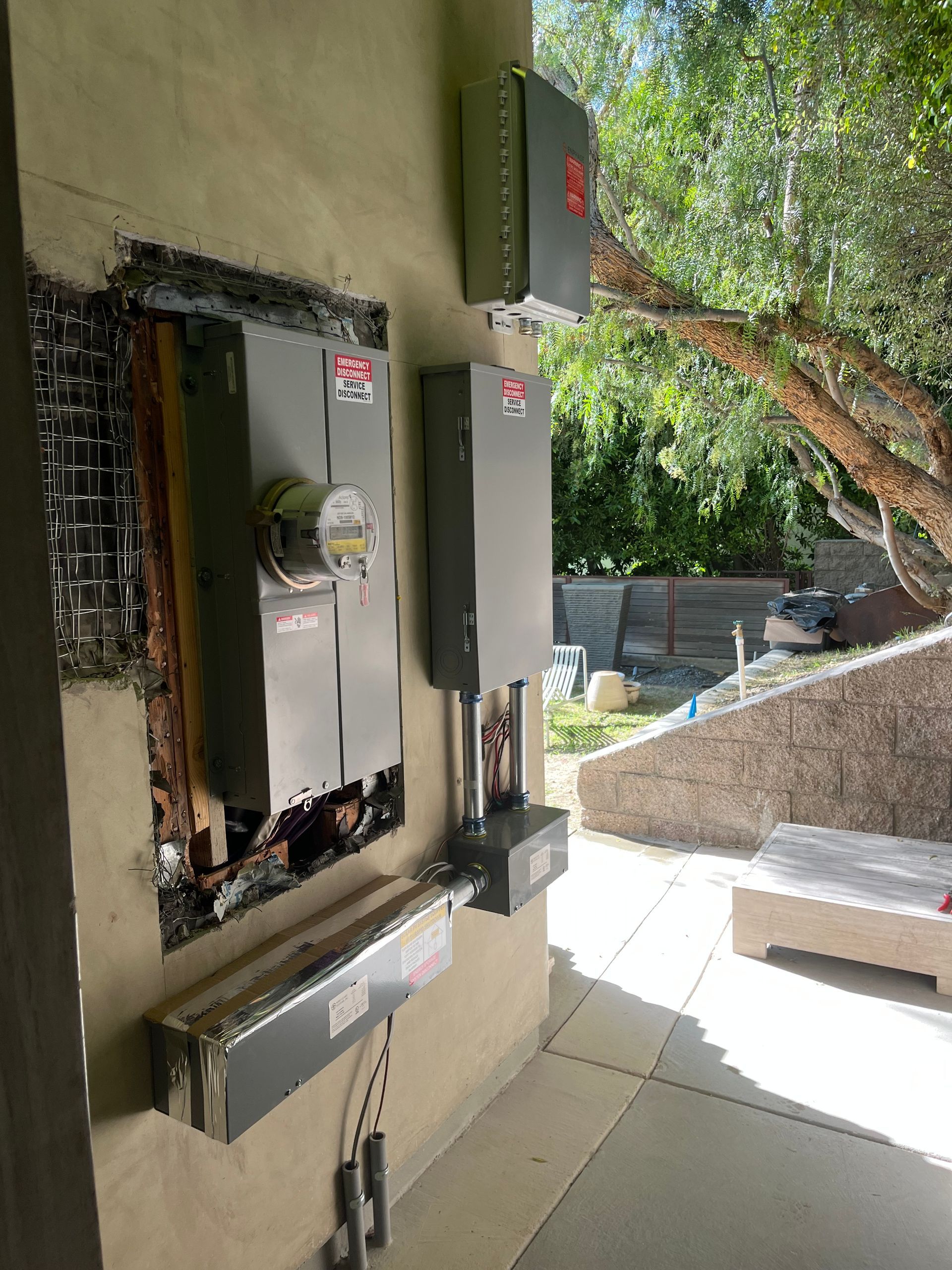Gray electrical meter panel and utility boxes mounted on an exterior stucco wall outdoors next to a landscaped area.