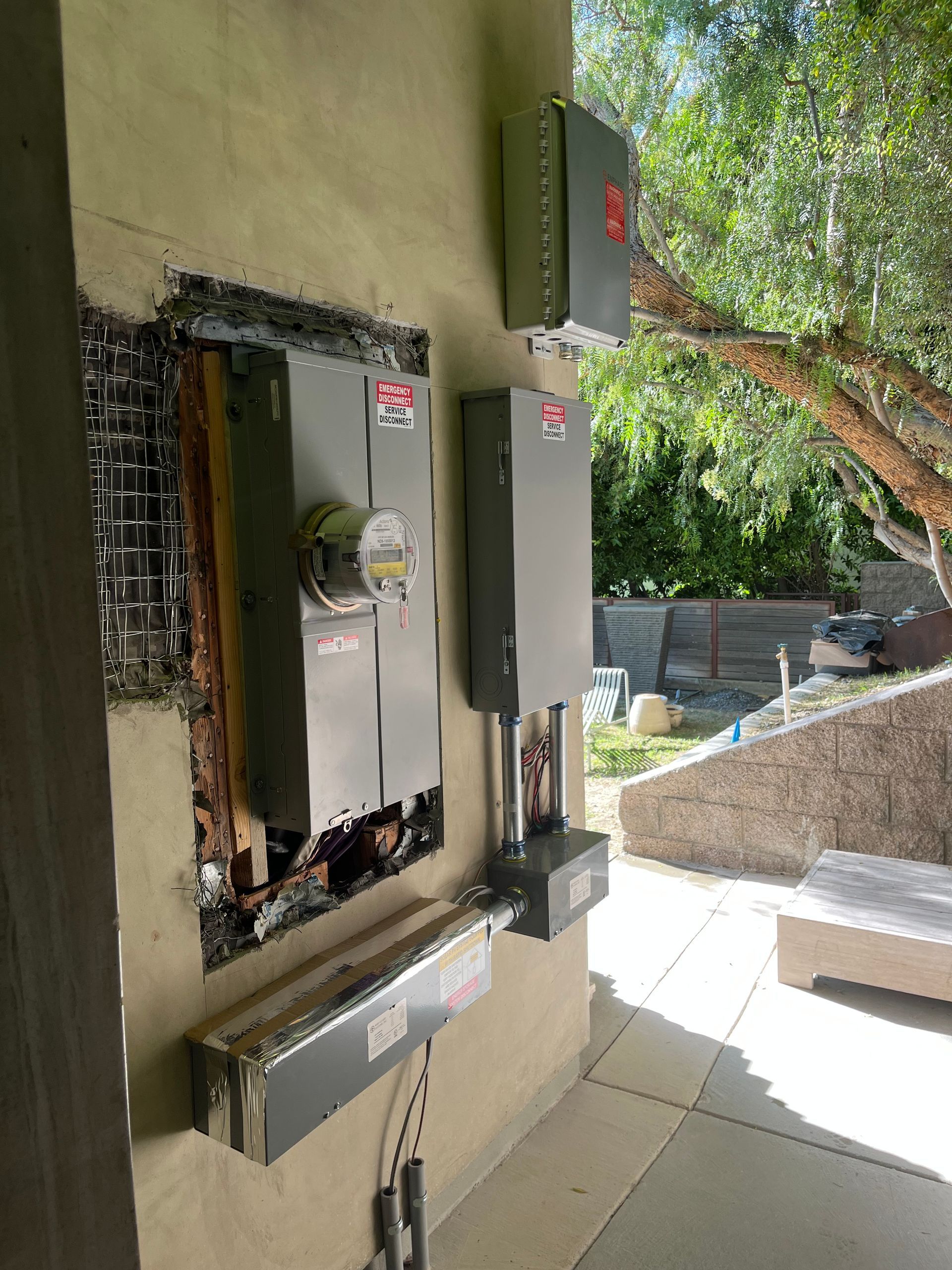 An electrical meter and service equipment boxes are mounted on an exterior stucco wall near an outdoor space.