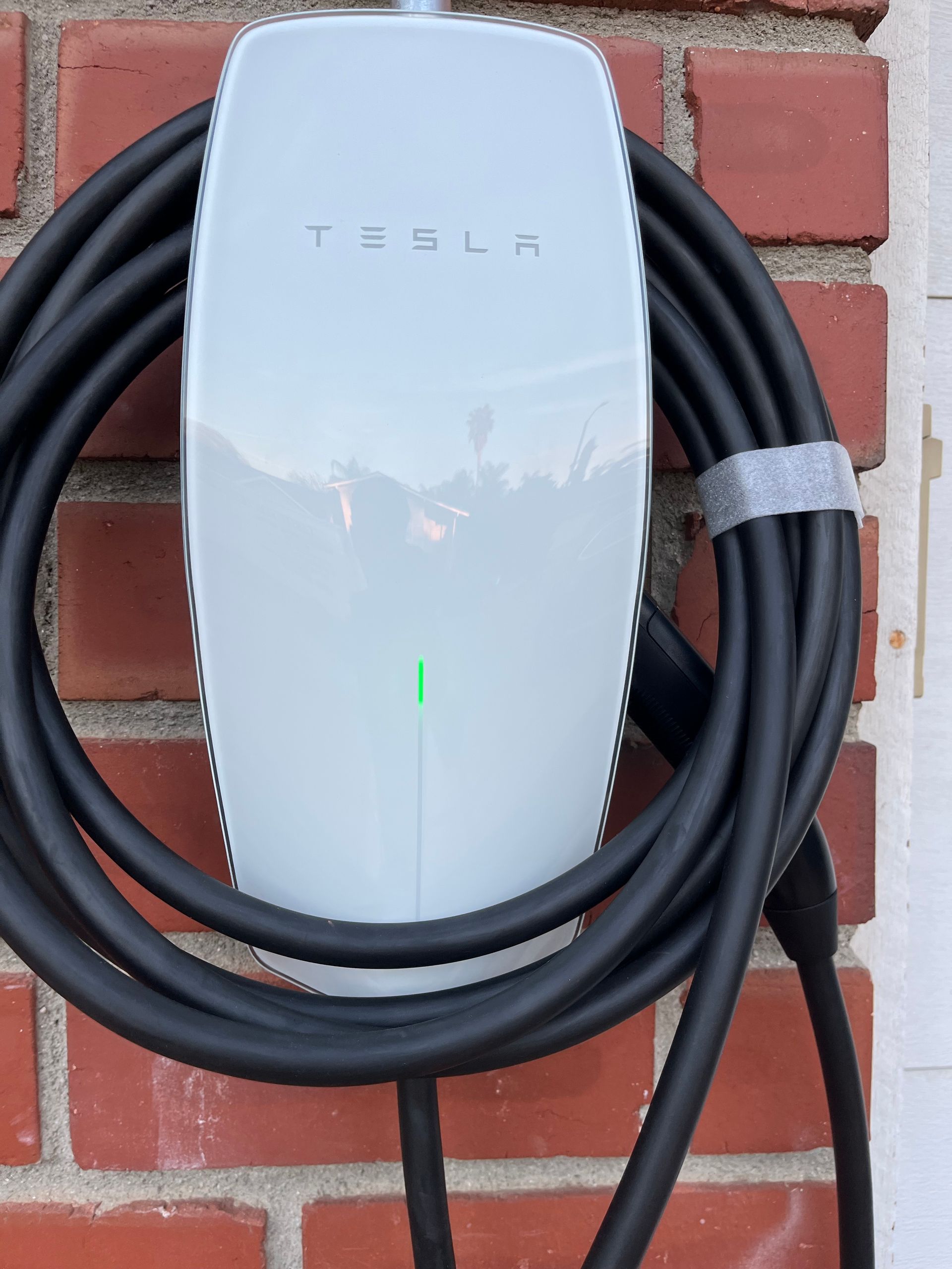 A white Tesla Wall Connector mounted on a brick wall with its black charging cable coiled around it, showing a green light.