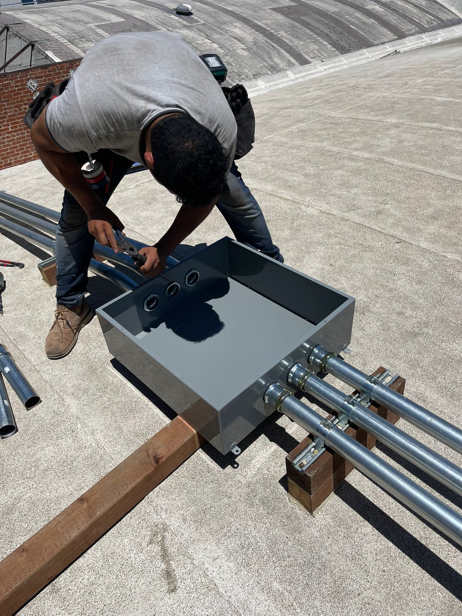 A construction worker installs metal conduits into a large electrical junction box on a flat gravel rooftop.