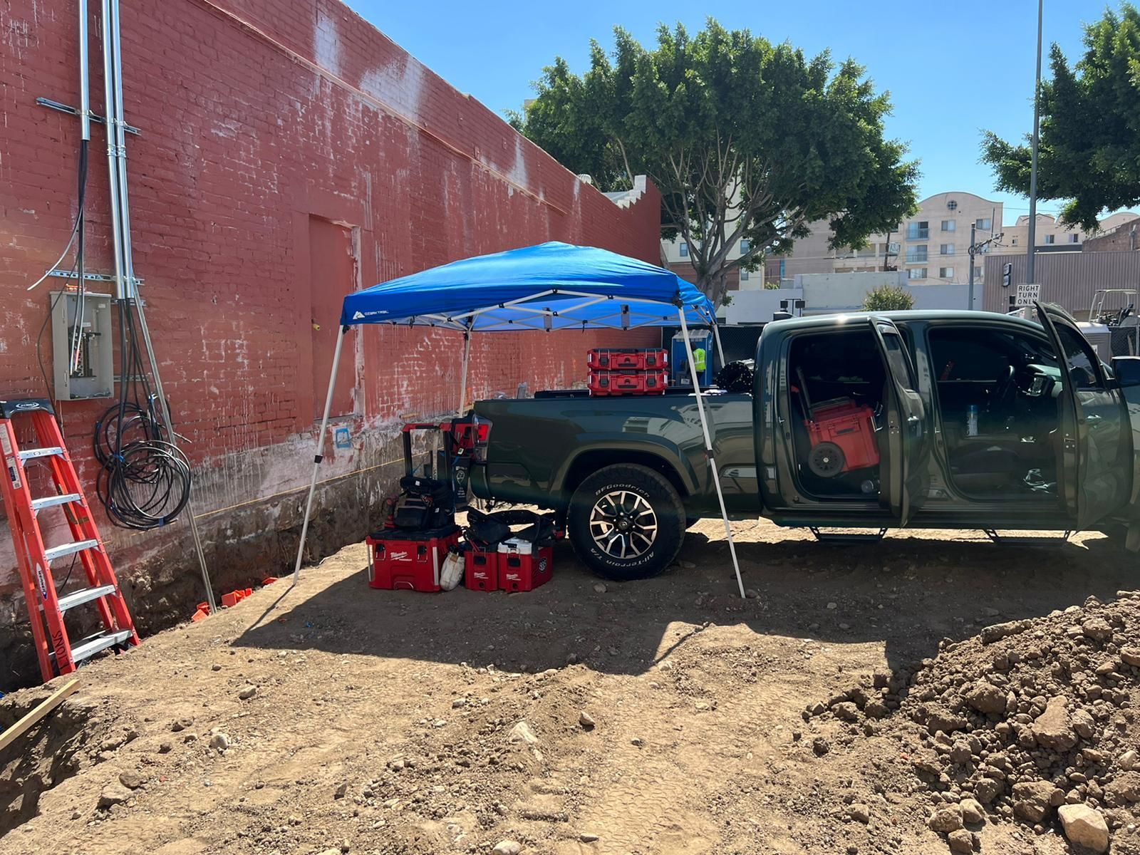 A dark green pickup truck parked on a dirt lot next to a brick wall, with a blue canopy covering tools and equipment.
