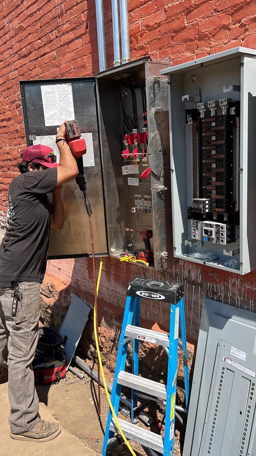 A worker using a power tool to install electrical equipment in metal boxes mounted on a brick wall.