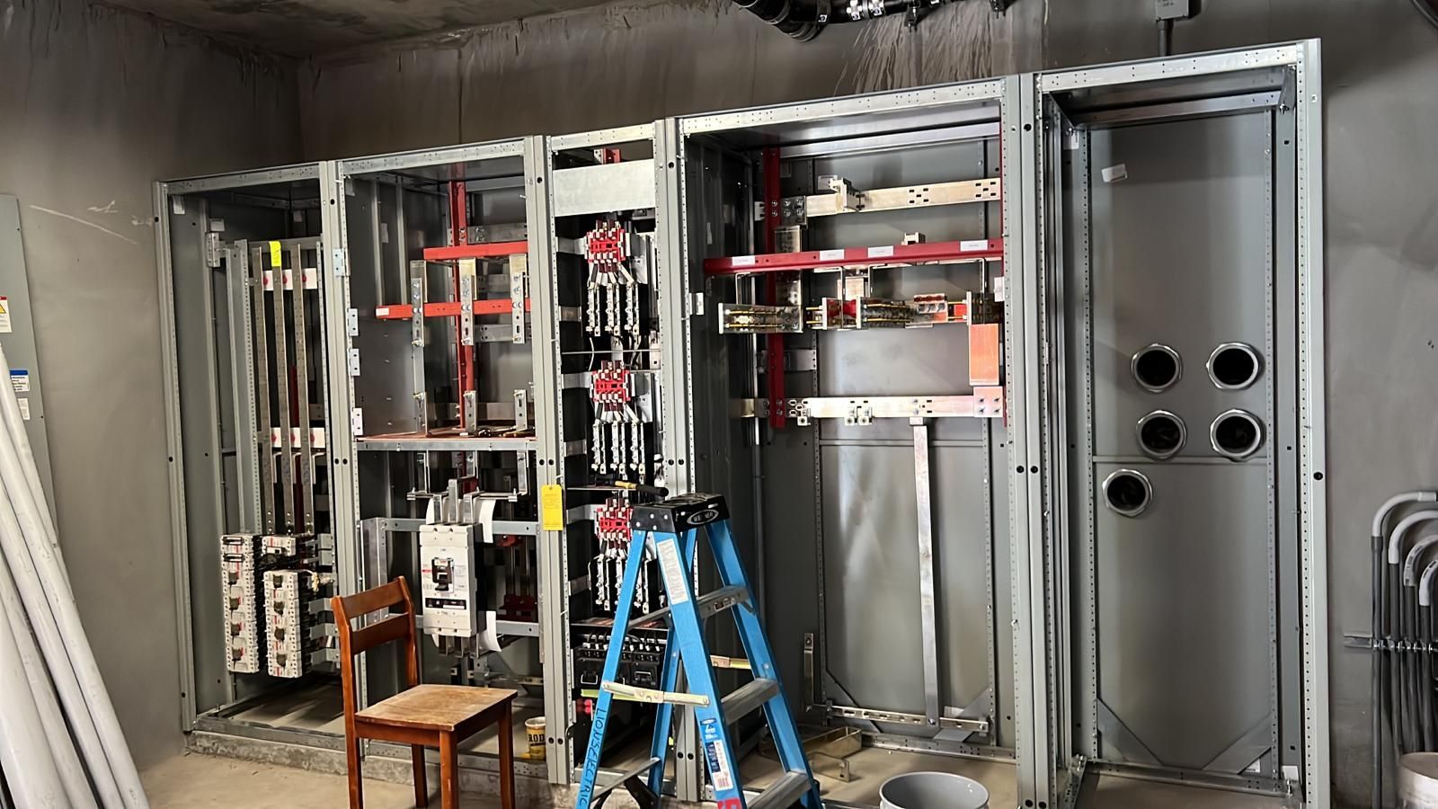 A large, open electrical distribution panel under installation in a concrete room, with a ladder and chair in front.
