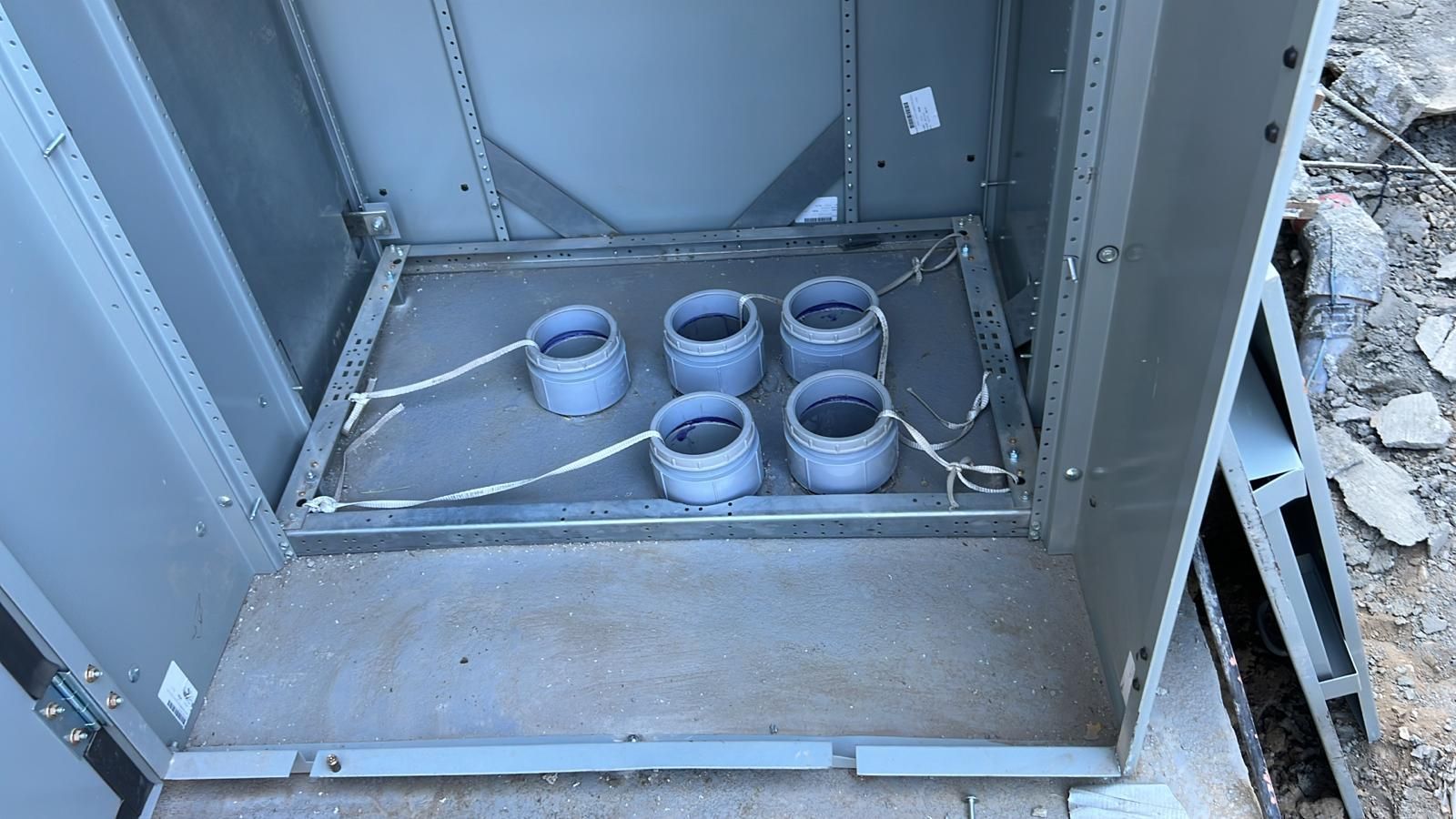 Five gray PVC pipe stubs installed on a metal frame inside a gray industrial enclosure box at an outdoor construction site.