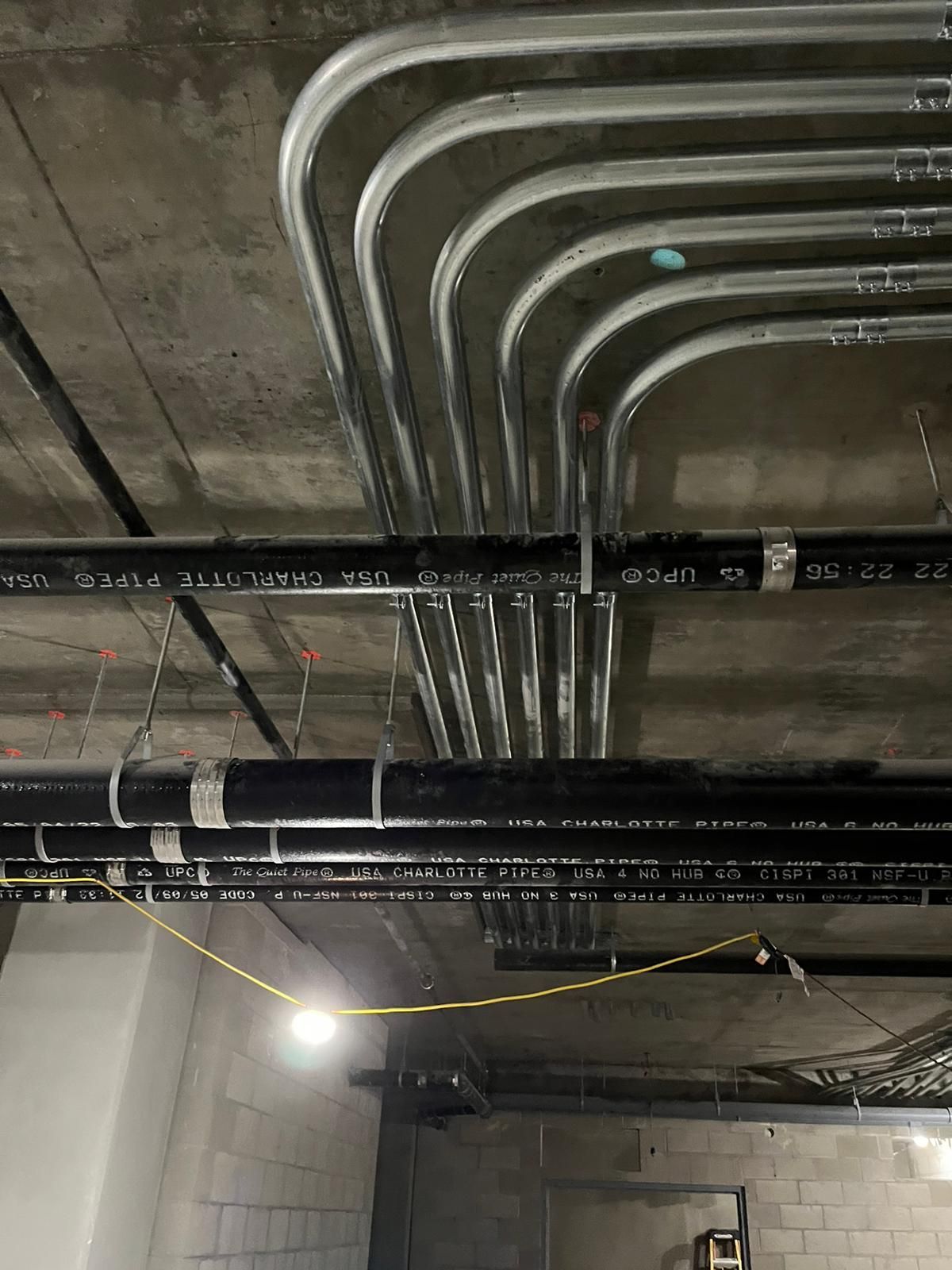 A series of parallel electrical conduits curve along a concrete ceiling, crossing above several horizontal black pipes.