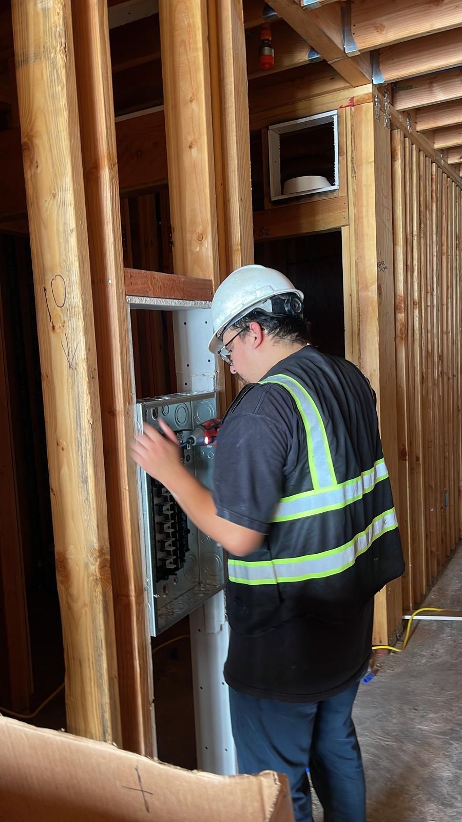 A person wearing a hard hat and high-visibility vest works on an electrical panel inside a wooden building frame.