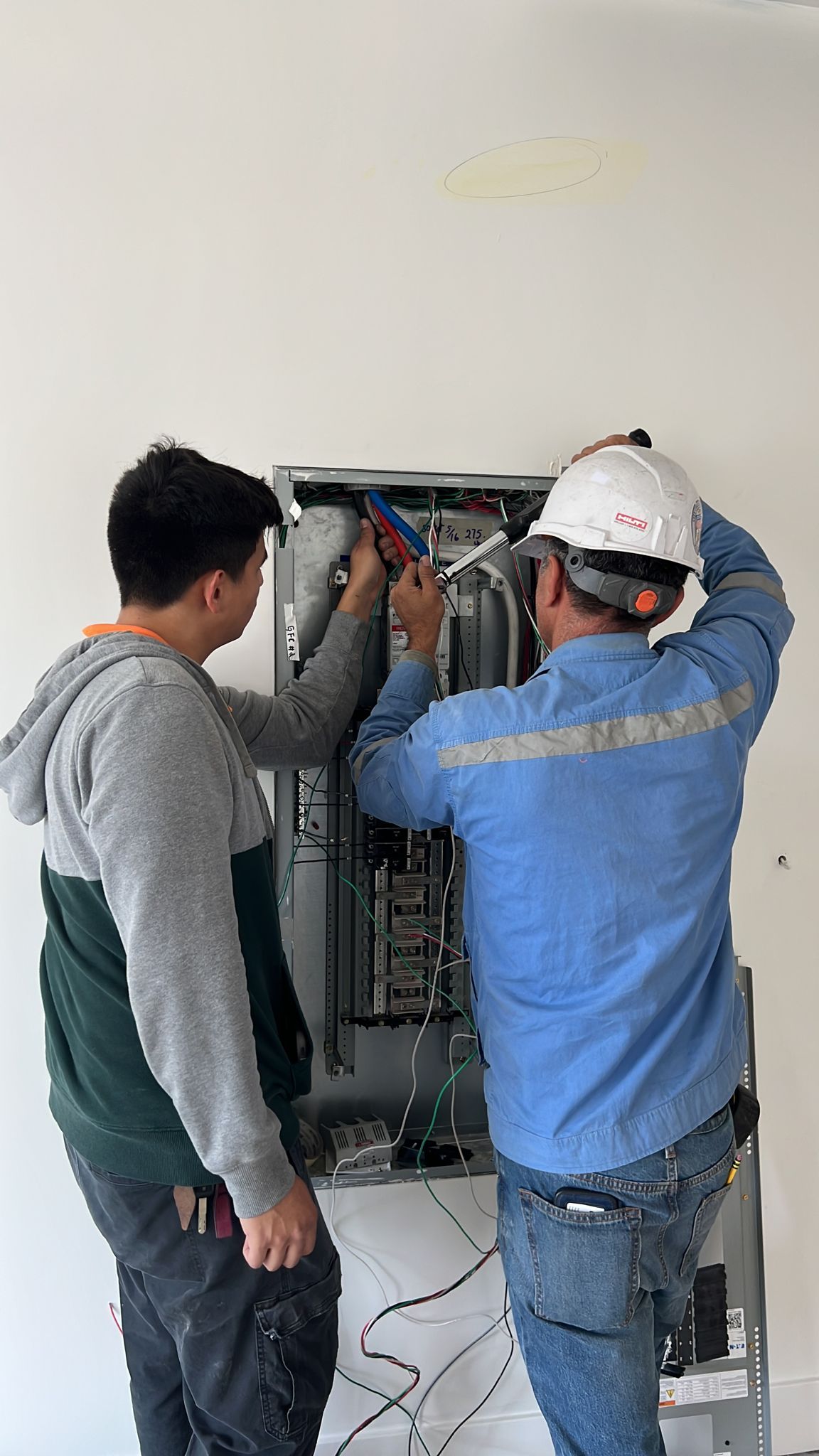 Two people work on the open interior of a wall-mounted electrical panel.
