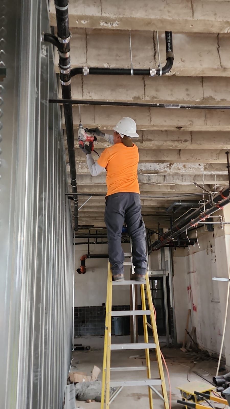 A construction worker in an orange shirt stands on a ladder, using a power tool to work on pipes near a concrete ceiling.