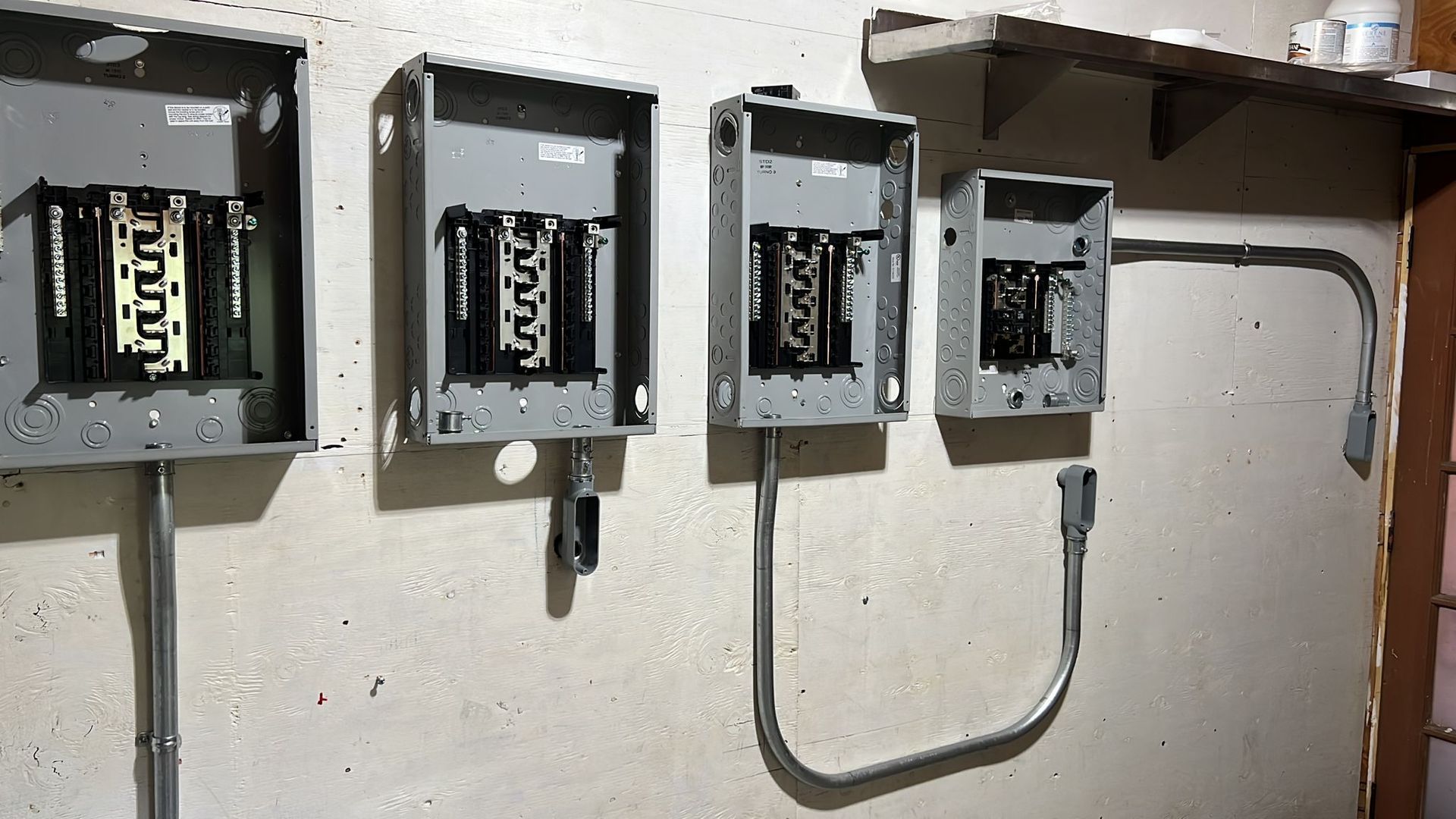 Four empty gray electrical breaker boxes mounted on a white wall with metal conduit piping connecting them.
