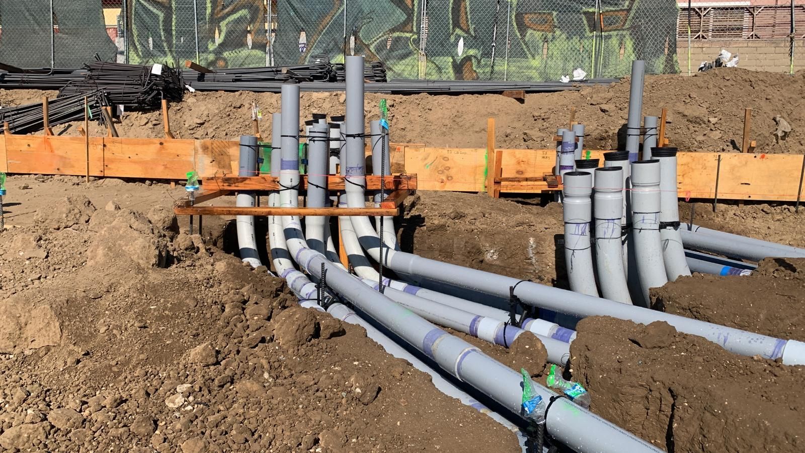 A construction site shows vertical grey PVC conduits emerging from trenches, braced by wooden boards in the dirt.