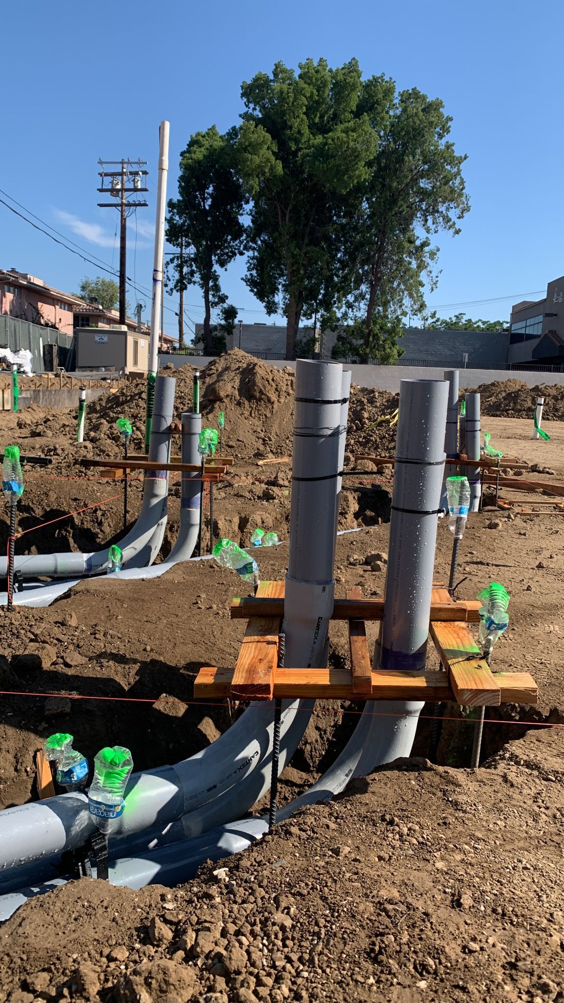 Vertical grey utility pipes rise from a dirt construction site, held in place by wooden supports under a clear blue sky.
