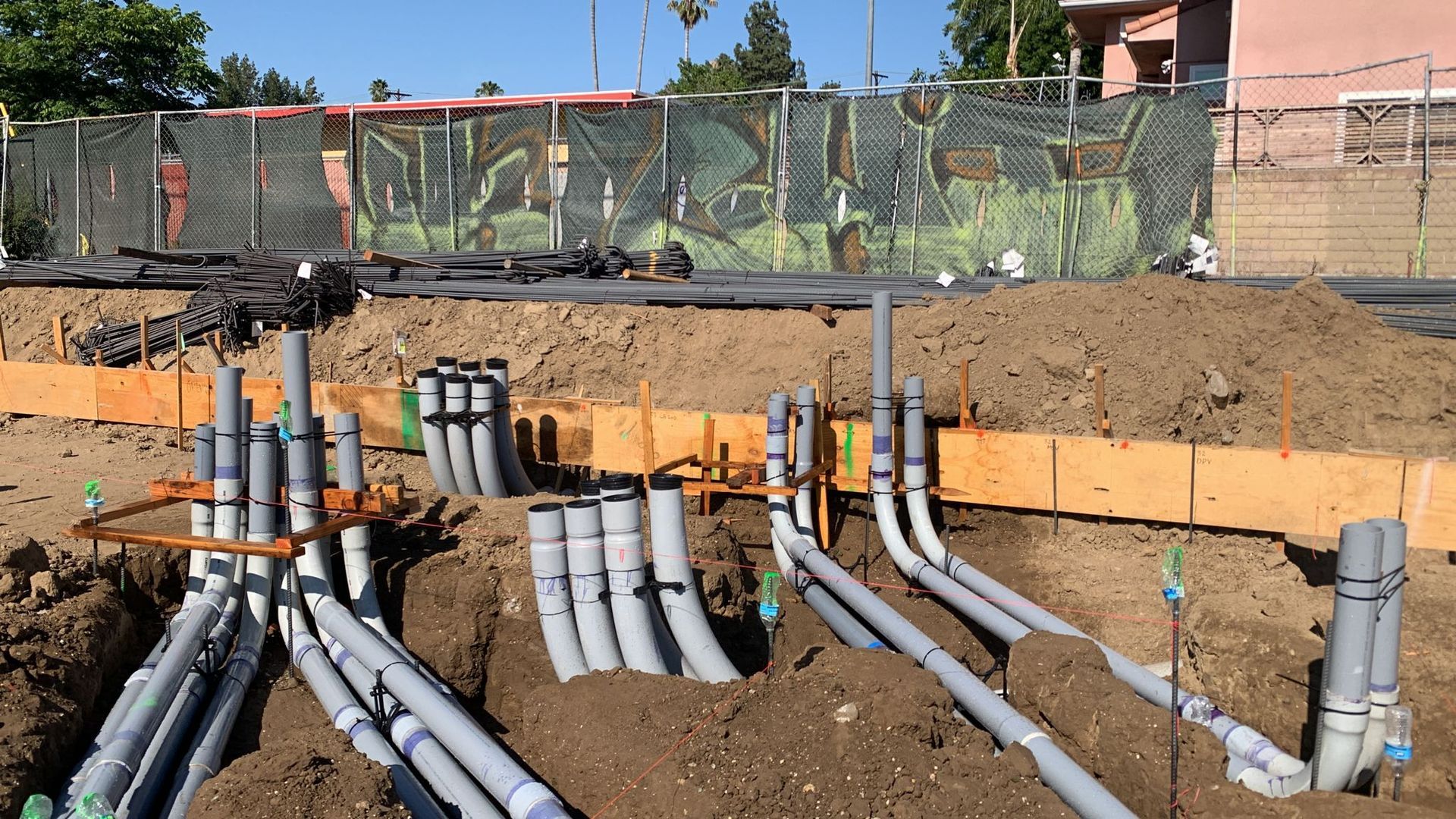 PVC electrical conduit pipes rising from a dirt construction site trench in front of wooden foundation forms.