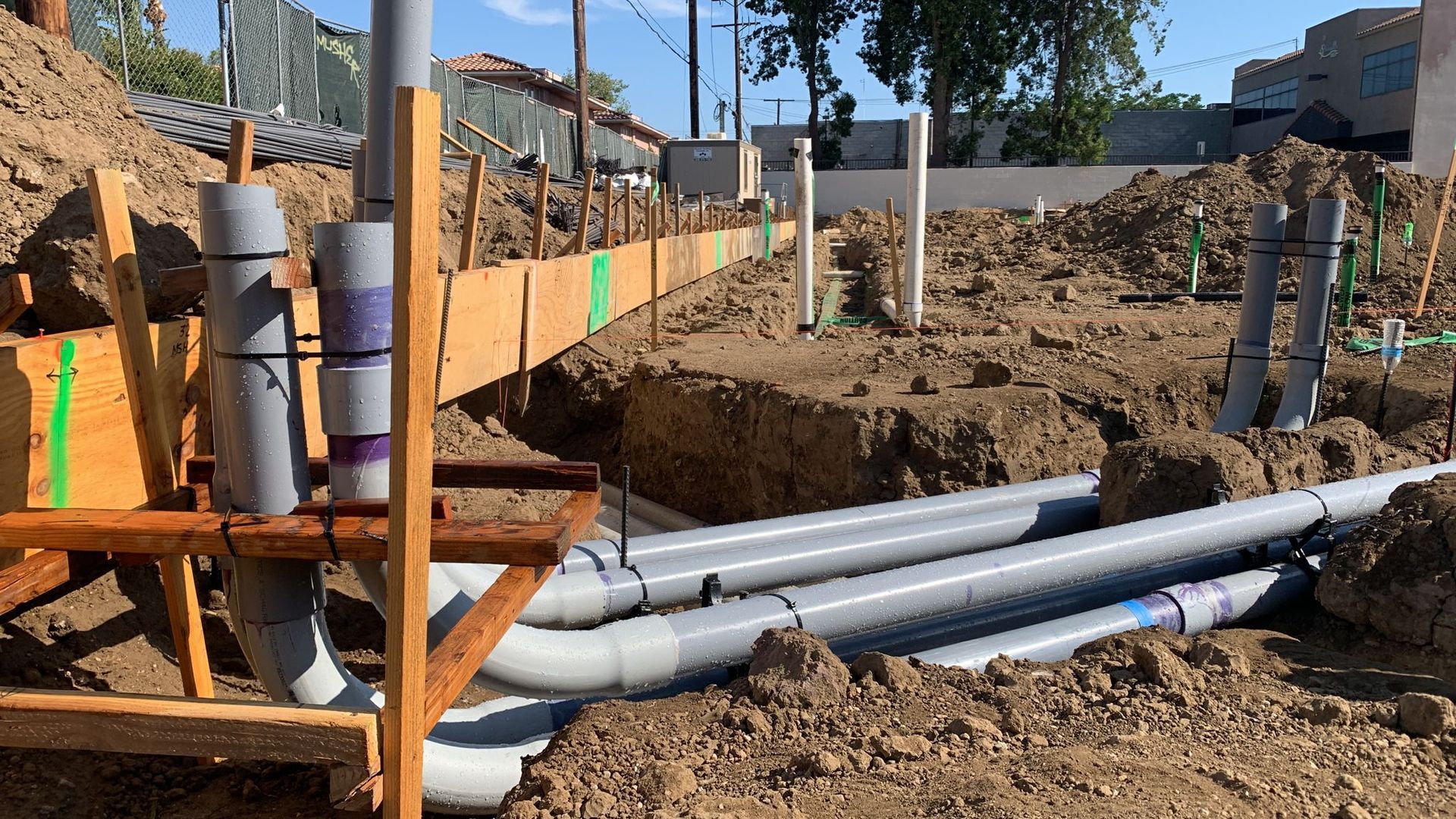 Construction site showing grey plumbing pipes installed in a dirt trench next to wooden foundation forms.