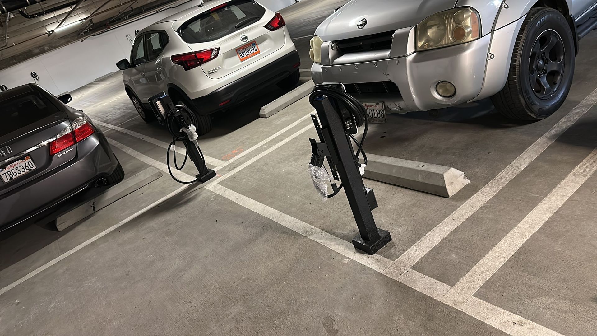 Two electric vehicle charging stations stand in a concrete parking garage between parked cars.