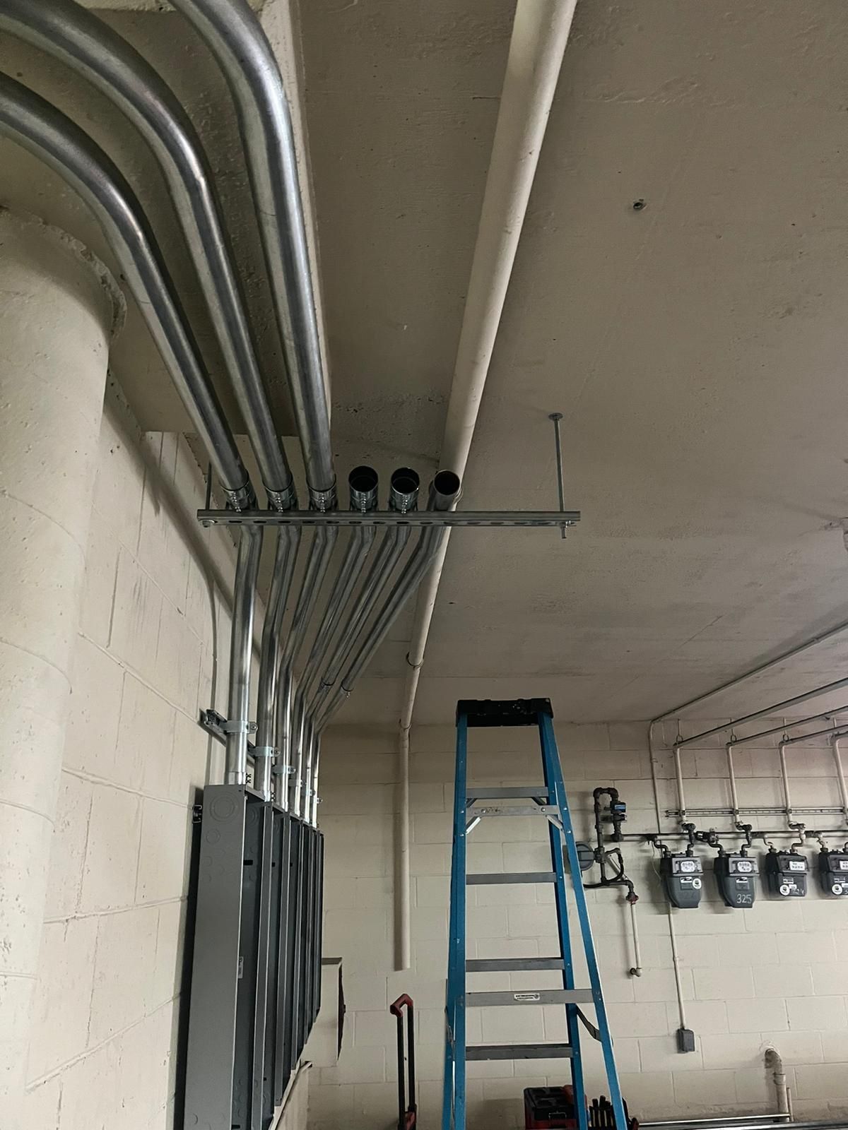 A blue ladder stands in a room with electrical conduits being installed along the ceiling above a row of power meters.