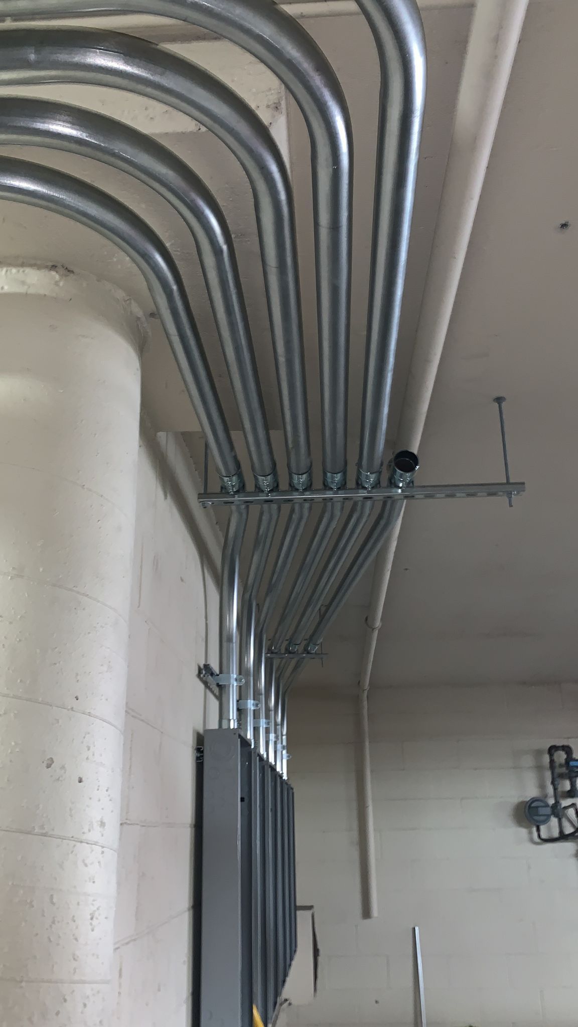 Five silver metal electrical conduits running along a beige wall and turning at a 90-degree angle to enter an electrical box.