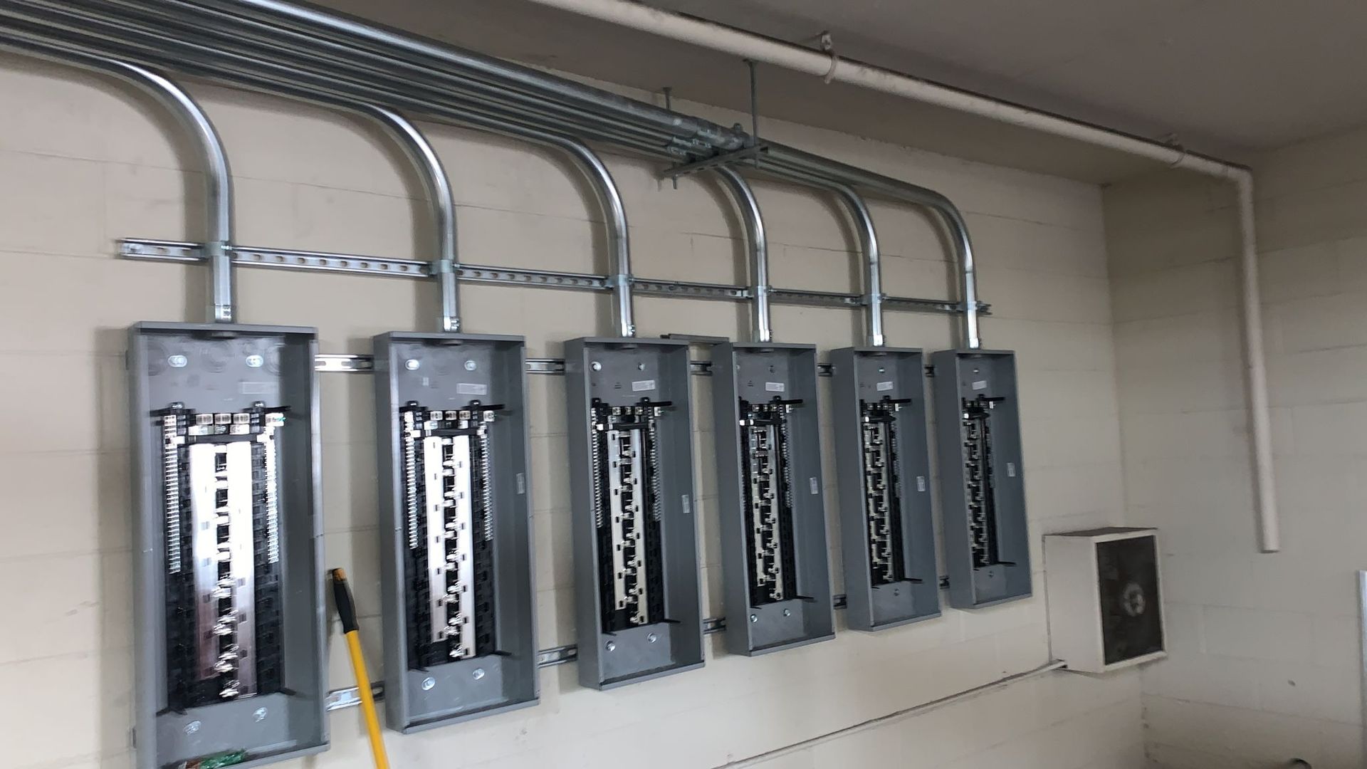Six electrical panel boxes mounted on a white wall, connected by overhead conduits and supported by metal strut channels.
