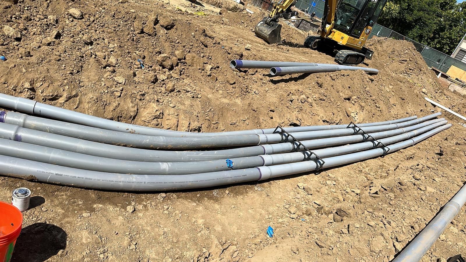 A view of several gray electrical conduits laid in a trench at a construction site, with a yellow excavator in the back.