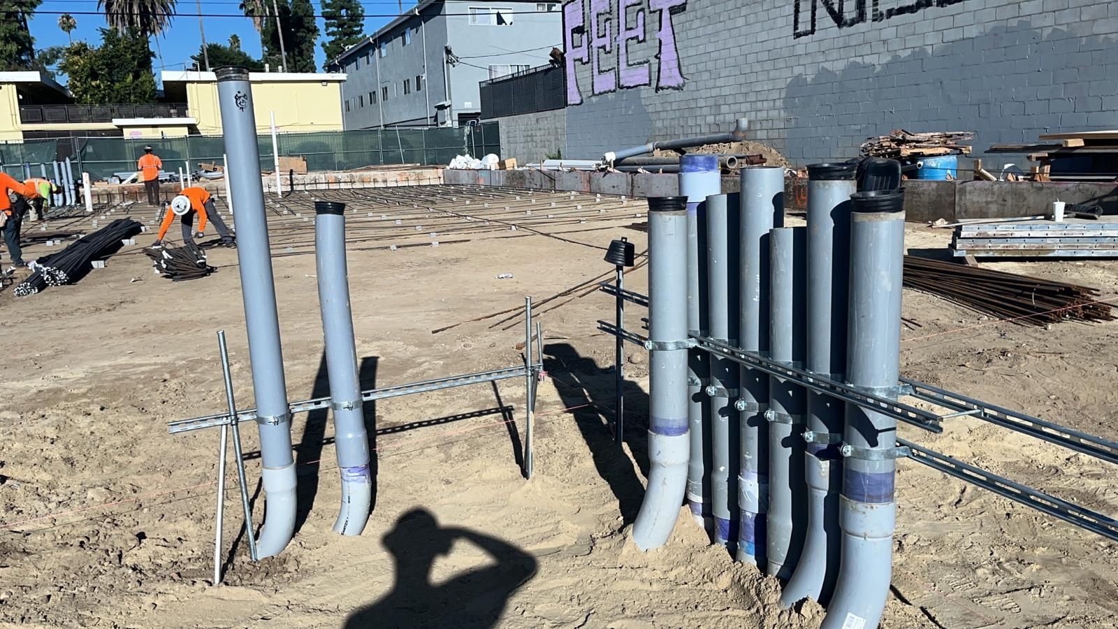 Grey conduits stand upright in a dirt construction site, supported by a metal frame with workers in the background.