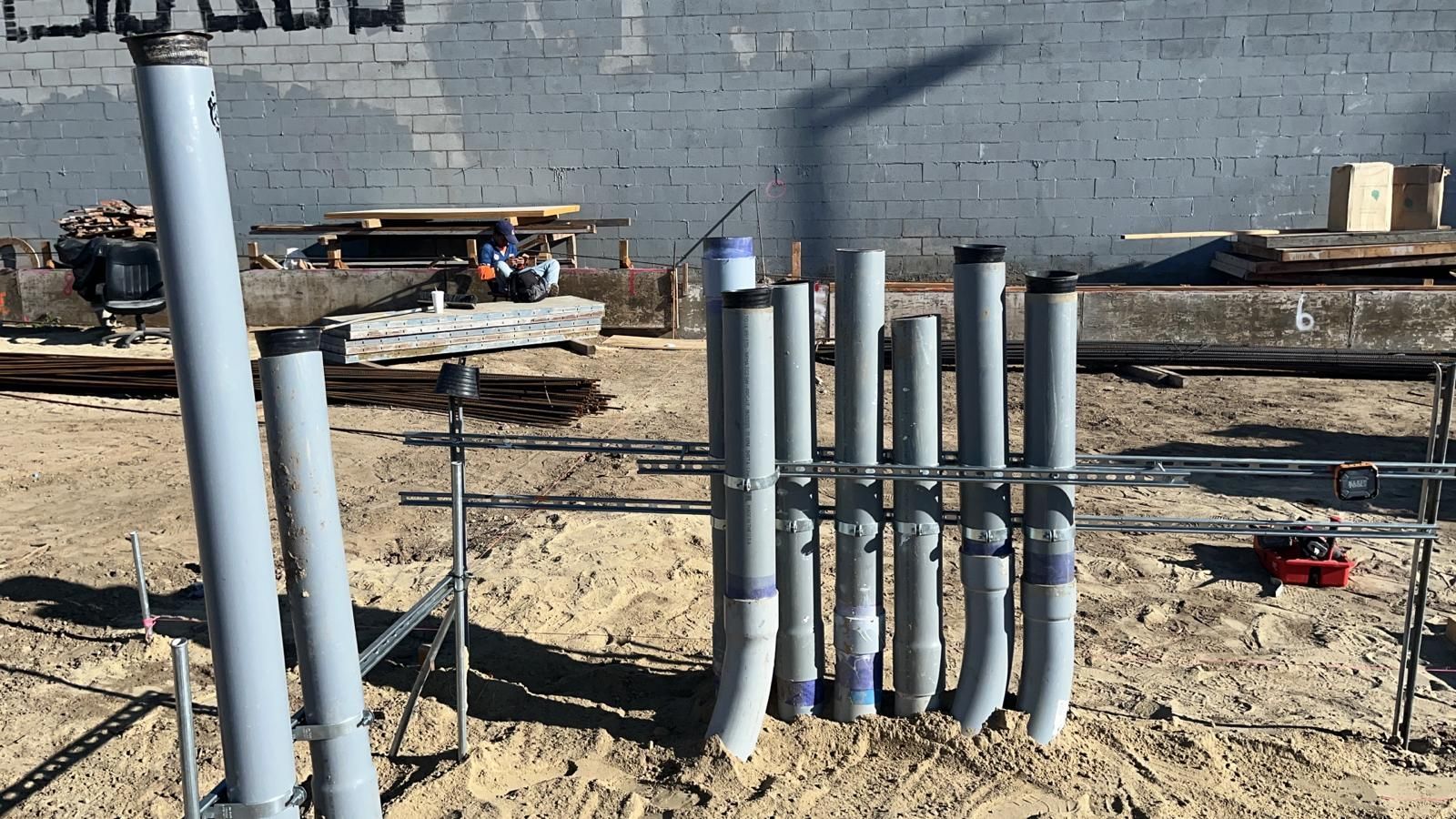 Several grey electrical conduits stand vertically in sandy soil, held in place by a metal support frame at a construction site.