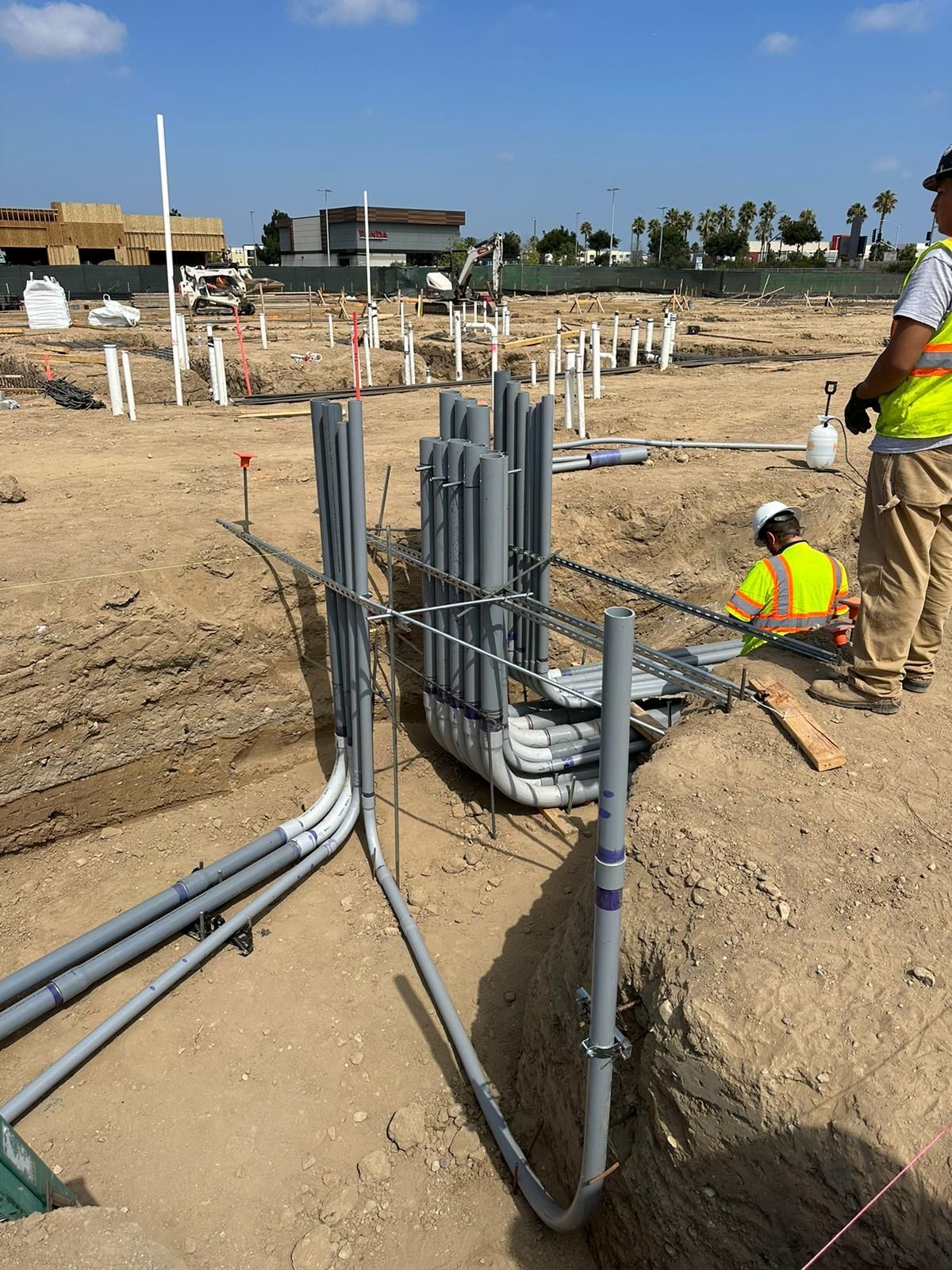 A construction worker in a high-visibility vest inspects electrical conduits emerging from a trench at a building site.