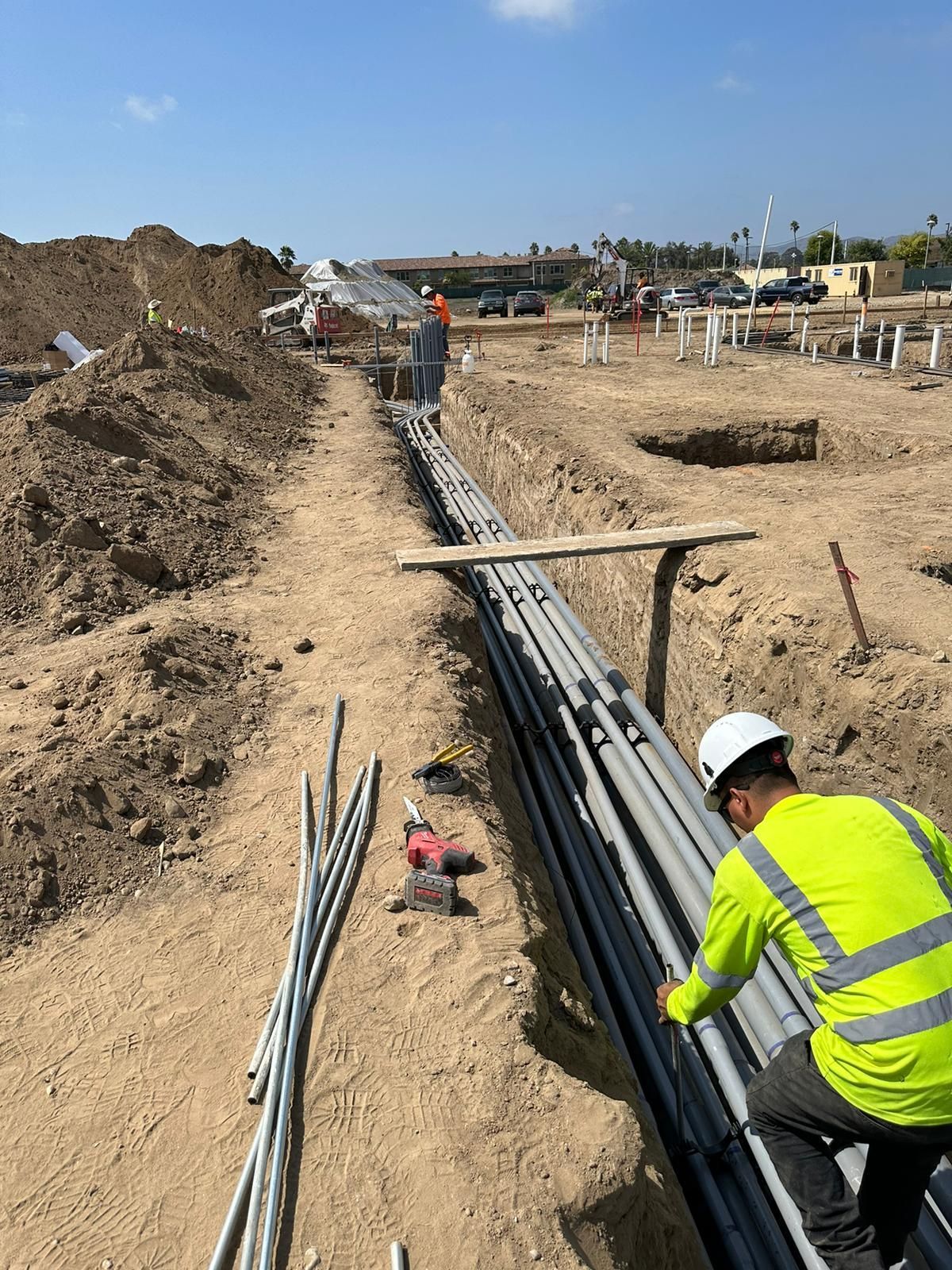 A worker in a high-visibility yellow shirt and hard hat installs multiple grey conduits in a trench at a construction site.