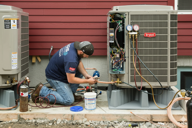 HVAC technician kneels, connecting copper tubes to an AC unit outside a red building.