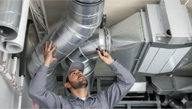 HVAC technician inspecting ductwork with a flashlight in a mechanical room, wearing a gray uniform.