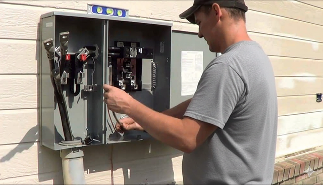 Man working on an electrical panel outside a building; he wears a cap and is smiling.
