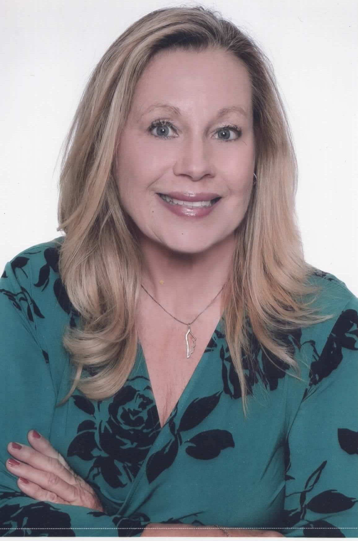 A smiling person with shoulder-length blonde hair, wearing a green floral top with arms crossed, against a white background.