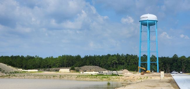 A light blue water tower stands tall near a body of water, a construction site, and a tree line under a cloudy sky.