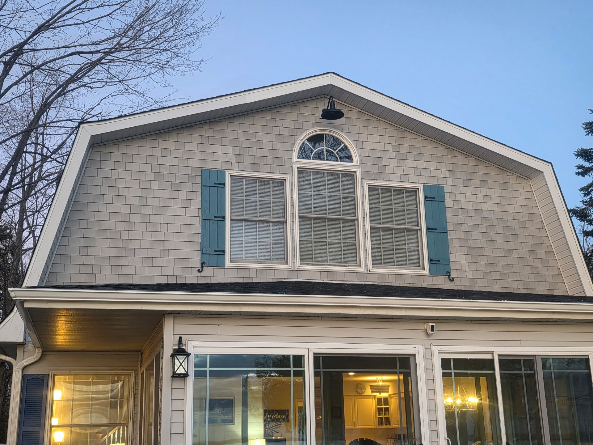A gambrel-roofed house with grey shingle siding, featuring a central arched window installed by Alpine Brothers Construction
