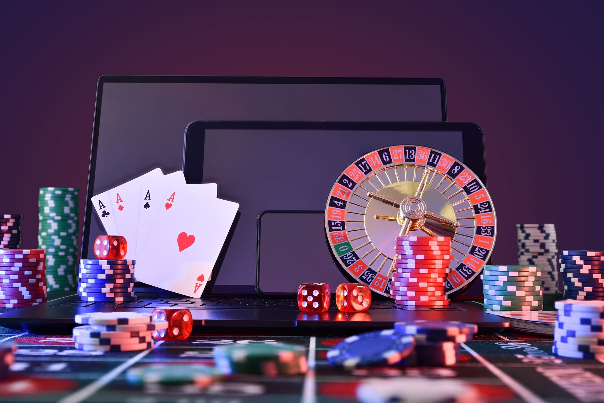 A laptop , tablet , roulette wheel , dice , playing cards and poker chips on a table