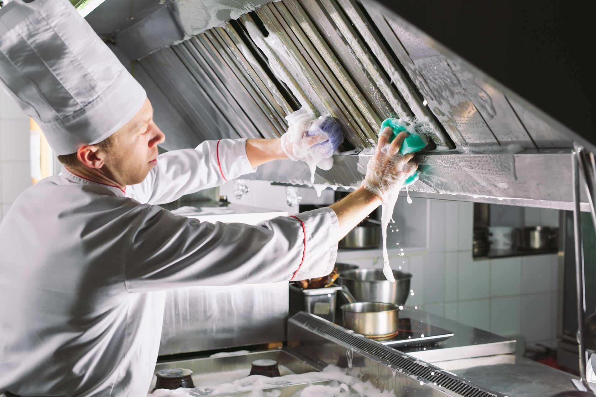 Chef cleaning grease from commercial kitchen hood using spray and sponge in busy kitchen.