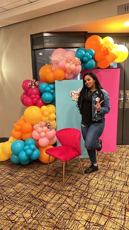Woman posing by colorful balloon arch and backdrop with a pink chair.