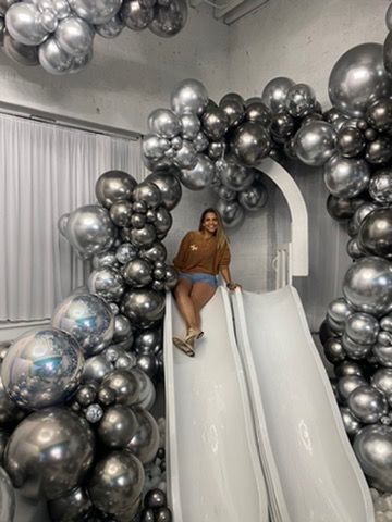 Woman on a white slide surrounded by silver and black balloons, inside a room.