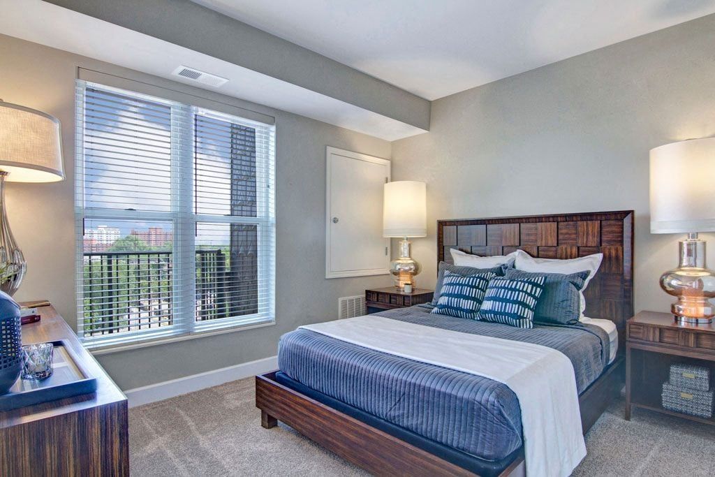 A bedroom with a bed , dresser , lamps , and two windows.
