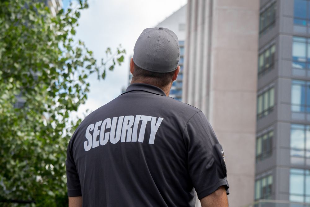 A man wearing a security shirt is standing in front of a building.