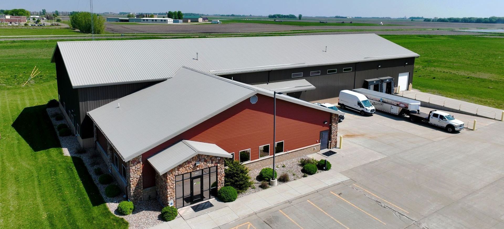 Aerial view of a commercial building with a red and stone facade, long gray warehouse, and parked trucks on a sunny day.