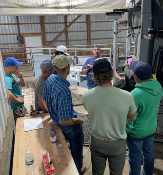 A group of men in a barn listens to a man speaking. The scene is indoors with agricultural equipment visible.