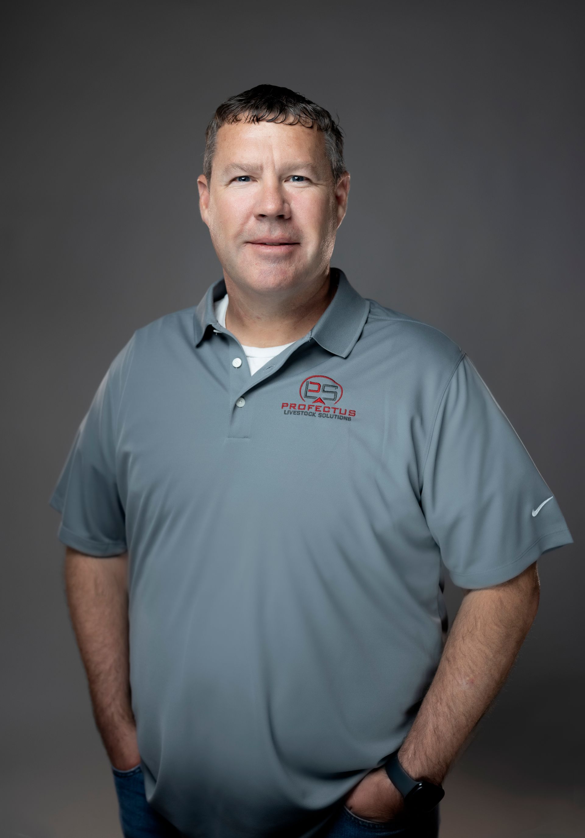 Man in a gray polo shirt with a logo, standing with hands in pockets against a gray background.