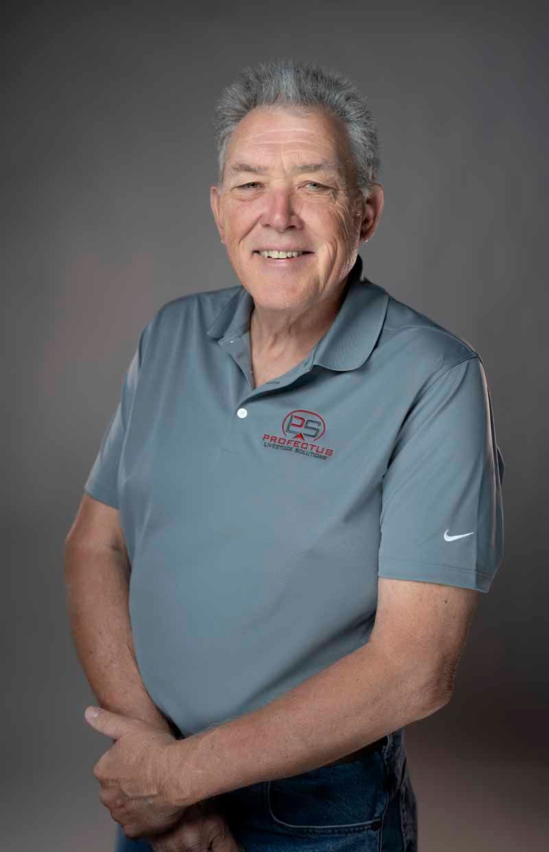 A smiling older man wearing a gray polo shirt with a logo and dark jeans stands in front of a gray background.
