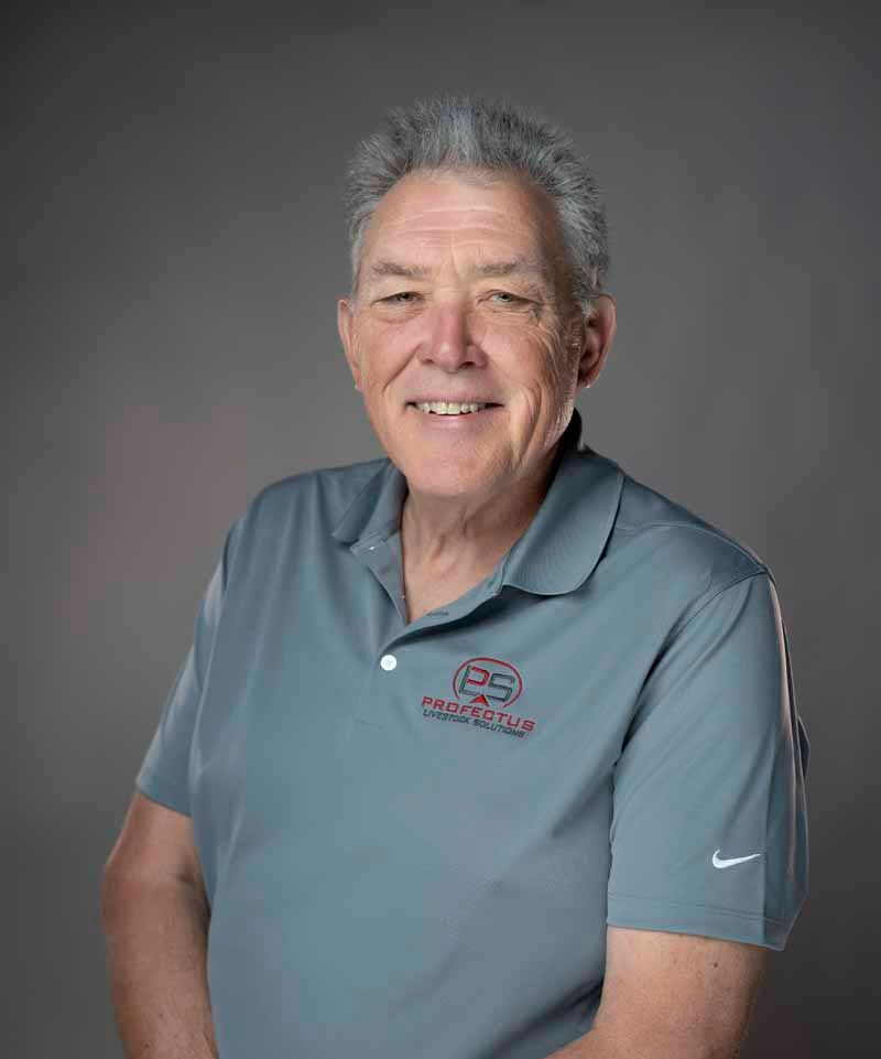 A smiling older man wearing a gray polo shirt with a logo and dark jeans stands in front of a gray background.