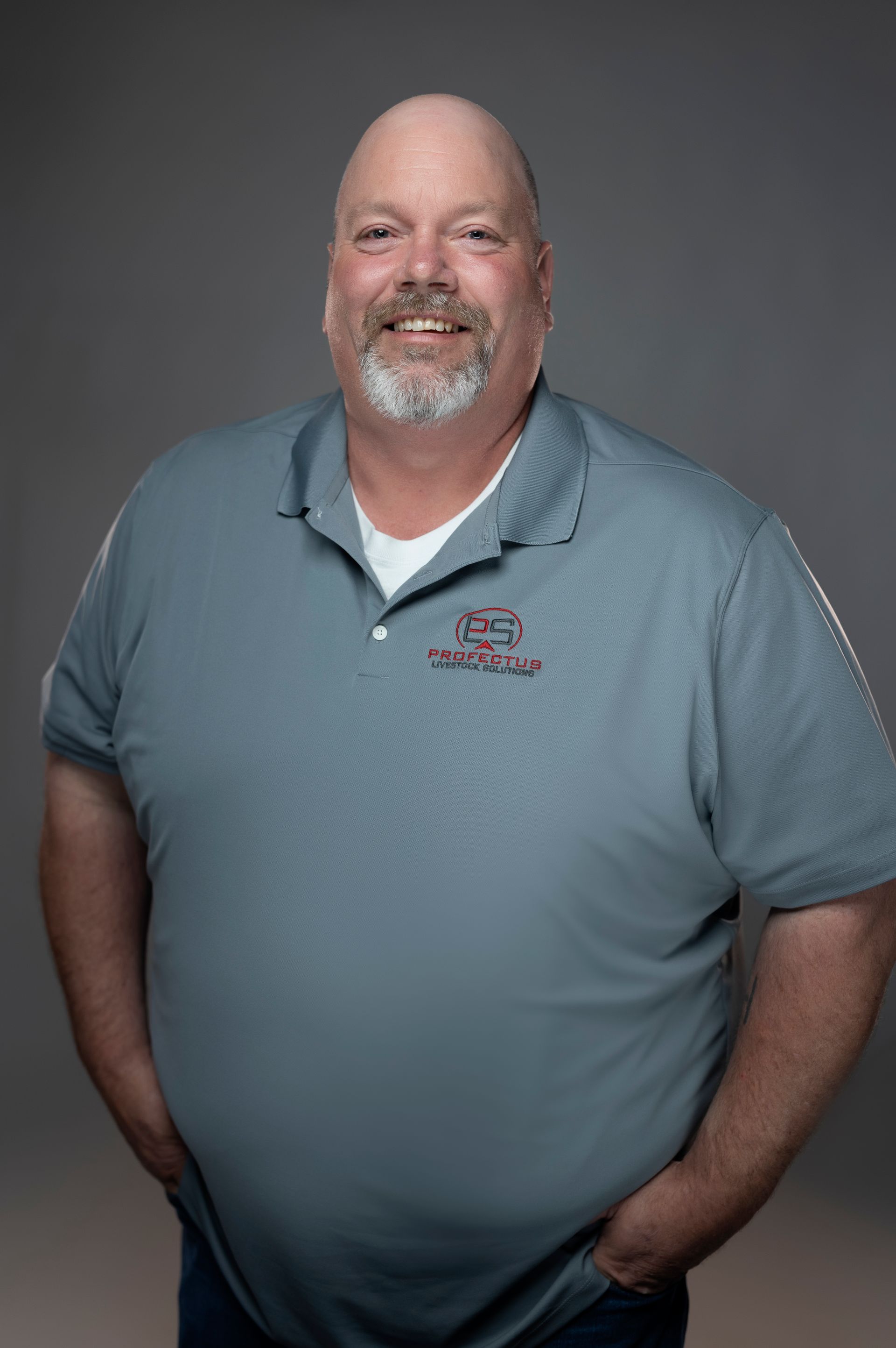 A smiling, balding man with a grey beard stands with his hands in his pockets, wearing a grey polo shirt.