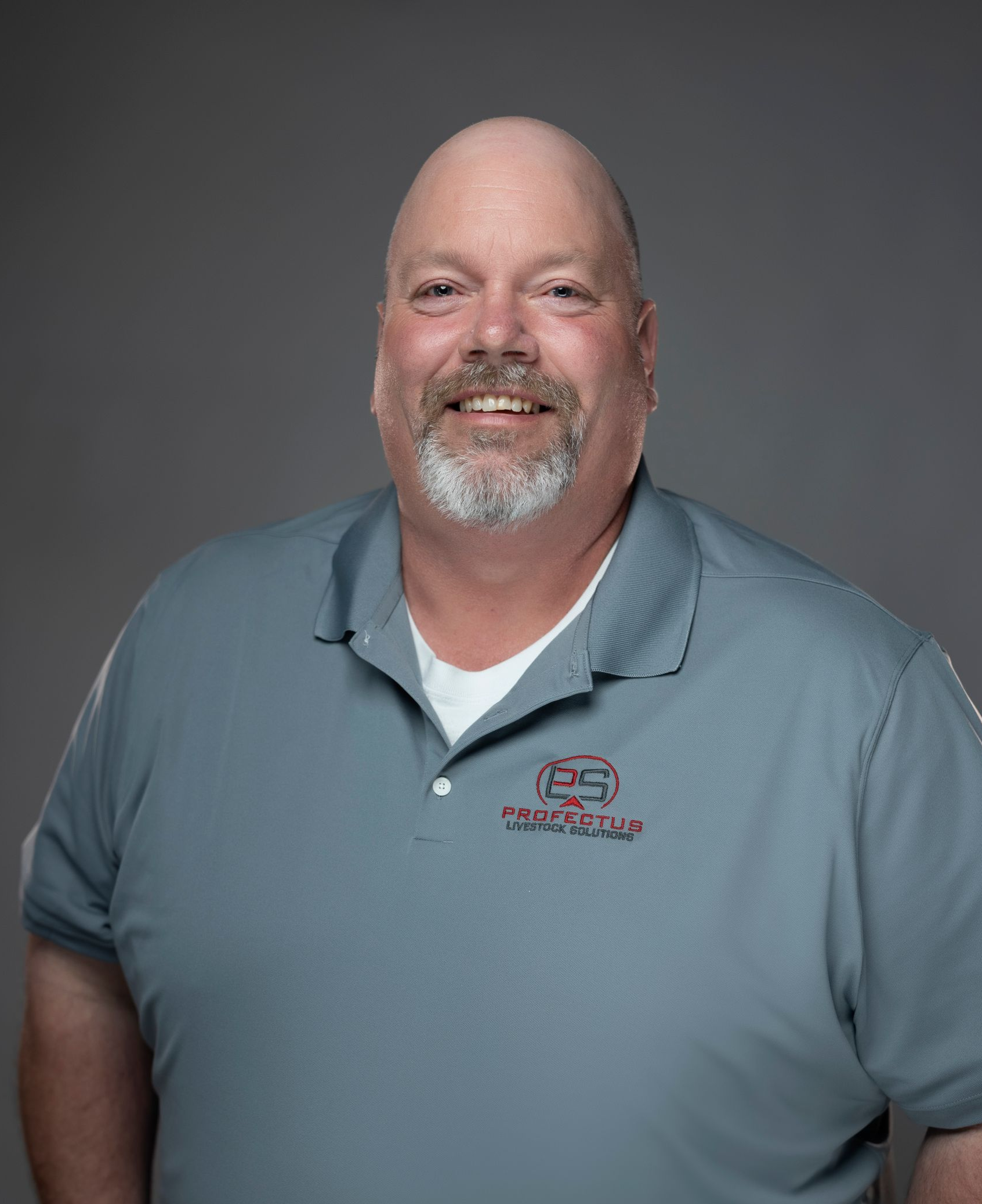 A smiling, balding man with a grey beard stands with his hands in his pockets, wearing a grey polo shirt.