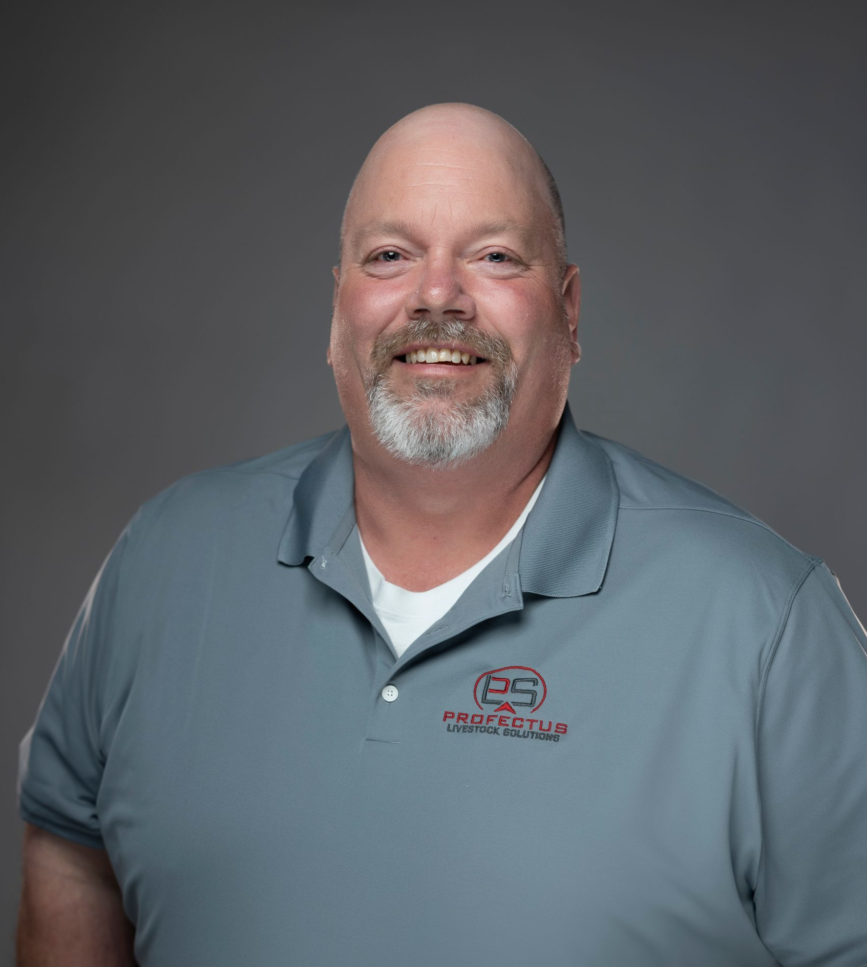 A smiling, balding man with a grey beard stands with his hands in his pockets, wearing a grey polo shirt.