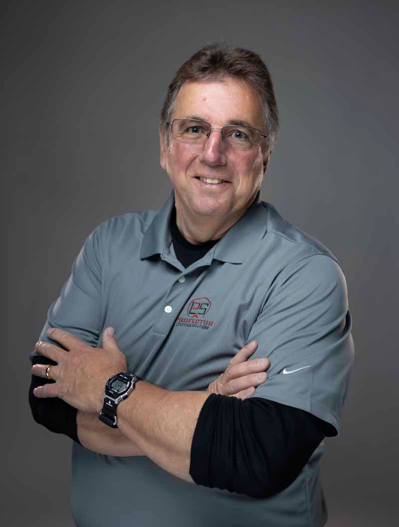 Man with glasses, arms crossed, wearing a gray polo shirt over a black shirt, smiling against a gray background.