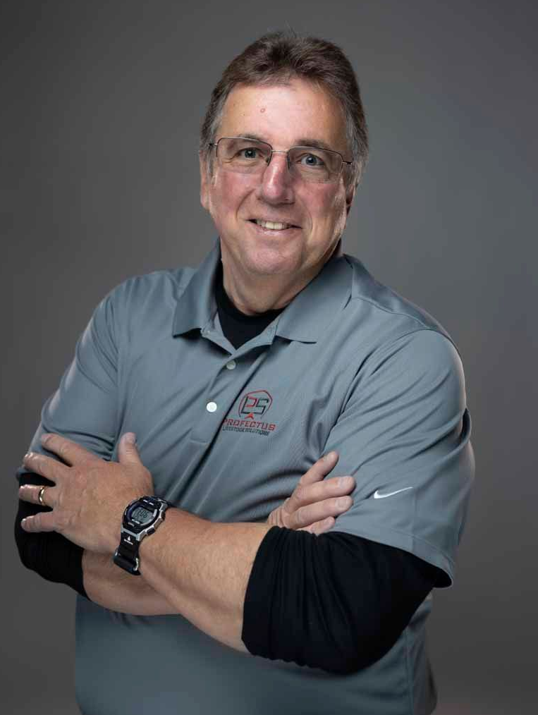 Man with glasses, arms crossed, wearing a gray polo shirt over a black shirt, smiling against a gray background.