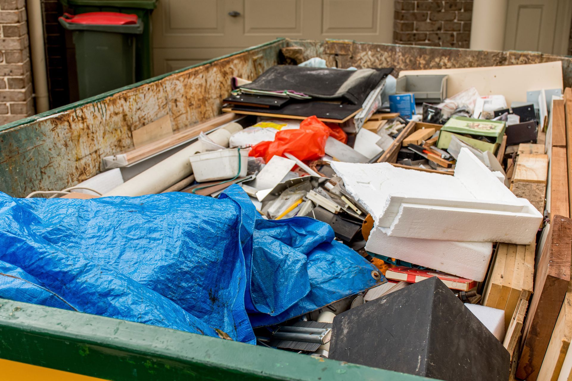 Overflowing dumpster filled with mixed construction debris, plastic sheeting, and foam boards